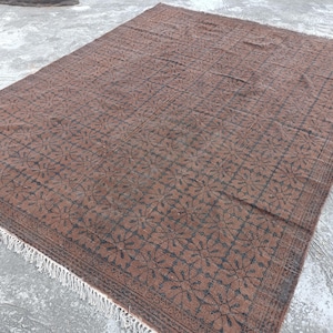 May include: A large, rectangular rug with a repeating floral pattern in shades of brown and black. The rug has a detailed border and a fringe along one edge. The rug is made of a woven material and is suitable for indoor use.