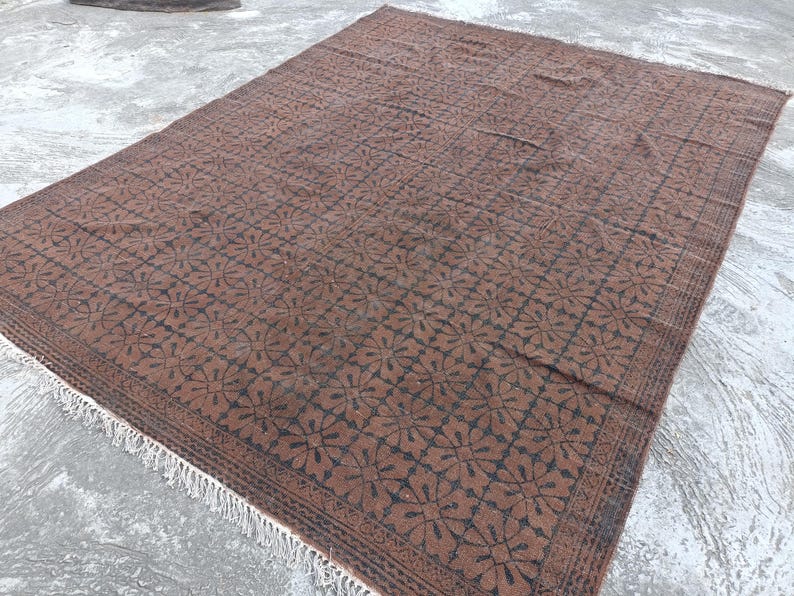 May include: A large, rectangular rug with a repeating floral pattern in shades of brown and black. The rug has a detailed border and a fringe along one edge. The rug is made of a woven material and is suitable for indoor use.