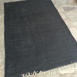 May include: A rectangular area rug in a deep charcoal black color featuring a visible woven texture throughout the surface. The rug is finished with short, light beige fringe detailing along the top and bottom edges, creating a contrast.
