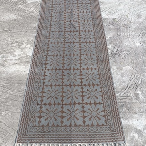 May include: A rectangular runner rug featuring a repeating floral pattern in brown and gray. The rug is bordered by a geometric zigzag design and finished with long, light-colored fringe tassels on both of the shorter ends of the textile.