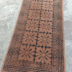 May include: A rectangular runner rug featuring a repeating black floral pattern on a terracotta orange background. The rug is framed by a black geometric border and finished with short, light-colored fringe tassels along the bottom edge.