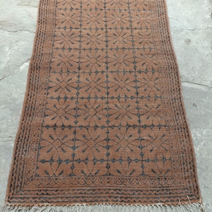 May include: A rectangular, brown rug with a repeating floral pattern in dark blue. The rug has a fringed edge and a detailed border. The rug is likely made of natural fibers and is suitable for a living room or bedroom.
