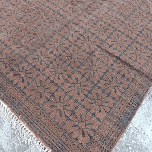 May include: Brown and black patterned rug with a floral design. The rug has a fringe on one edge and a border with a geometric pattern. The rug is made of a woven material.