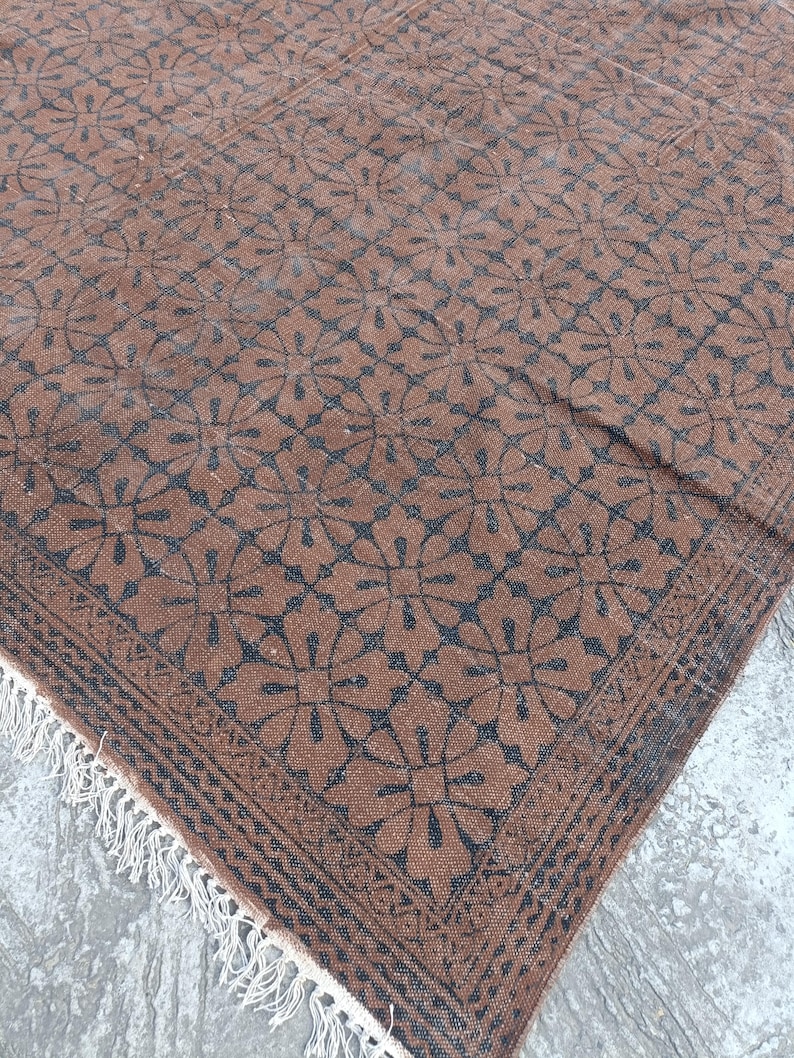 May include: Brown and black patterned rug with a floral design. The rug has a fringe on one edge and a border with a geometric pattern. The rug is made of a woven material.