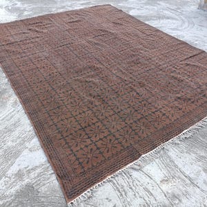 May include: A rectangular, brown rug with a repeating floral pattern in dark brown. The rug has a fringe along one edge and is placed on a concrete surface. The rug's design is symmetrical and detailed.