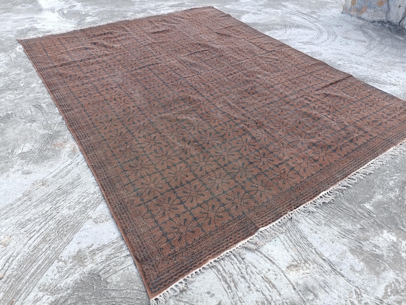 May include: A rectangular, brown rug with a repeating floral pattern in dark brown. The rug has a fringe along one edge and is placed on a concrete surface. The rug's design is symmetrical and detailed.
