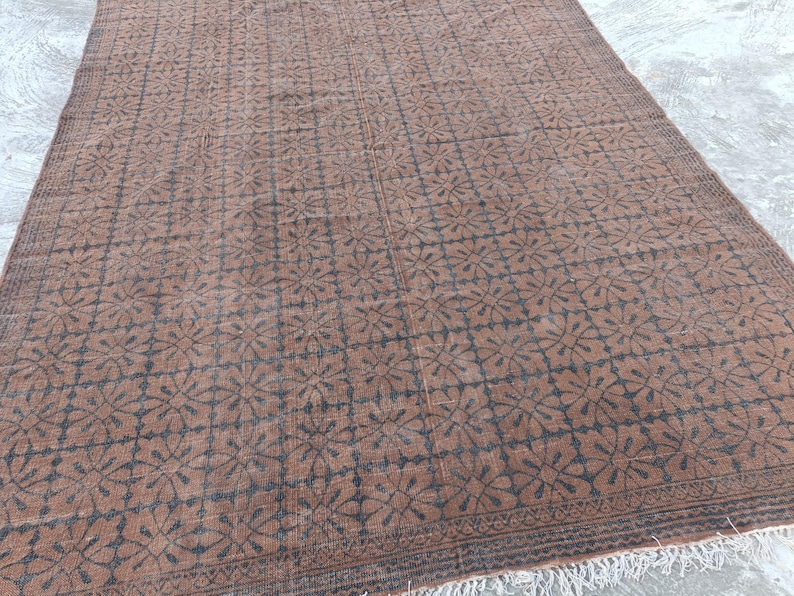 May include: A rectangular brown rug with a repeating floral pattern in dark brown. The rug has a fringe along the bottom edge and a decorative border. The rug is likely made of wool and is suitable for a living room or bedroom.