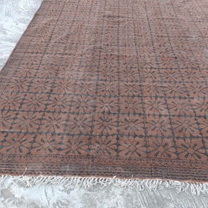 May include: A rectangular brown rug with a repeating floral pattern in dark brown. The rug has a fringe along the bottom edge. The pattern is a grid of stylized flowers.