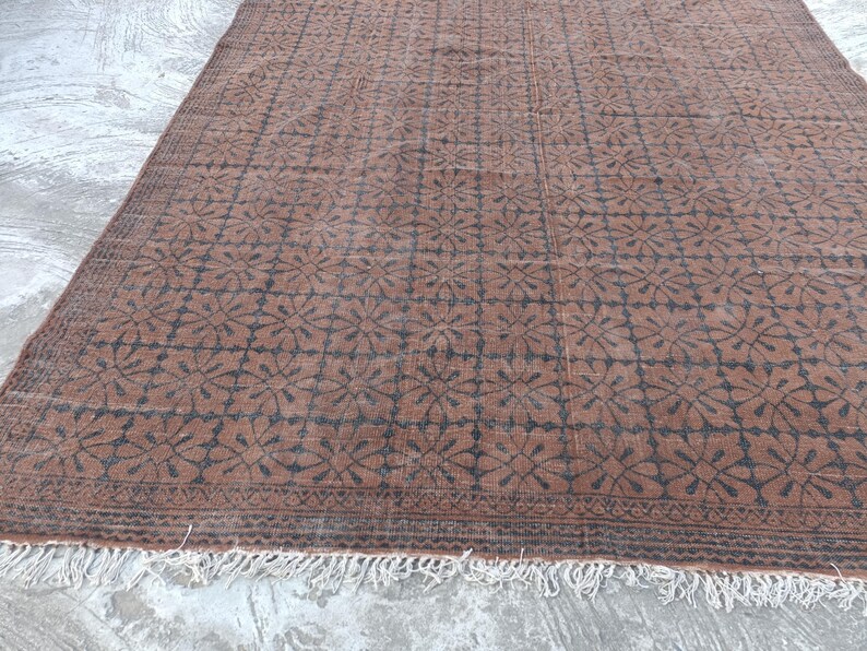 May include: A rectangular brown rug with a repeating floral pattern in dark brown. The rug has a fringe along the bottom edge. The pattern is a grid of stylized flowers.
