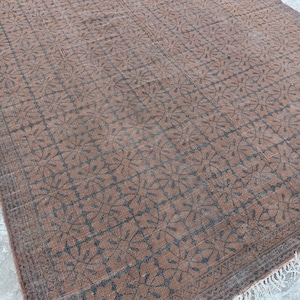 May include: A rectangular brown rug with a repeating floral pattern in dark gray. The rug has a fringe along one edge and is placed on a concrete surface. The rug's design is symmetrical and detailed.