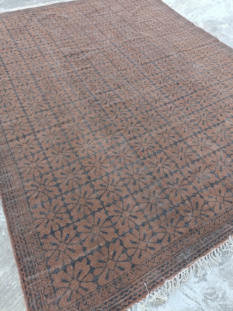 May include: A rectangular brown rug with a repeating floral pattern in dark gray. The rug has a fringe along one edge and is placed on a concrete surface. The rug's design is symmetrical and detailed.