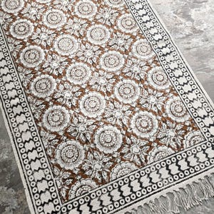 May include: A rectangular brown and white floral patterned rug with a black and white border and fringe. The rug features a repeating floral design in white against a brown background. The border has a geometric pattern.