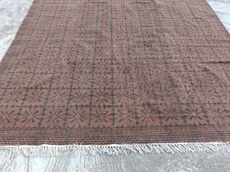 May include: Brown and black patterned rug with a floral design. The rug has a rectangular shape and a fringe along the bottom edge. The pattern is repeated across the surface.