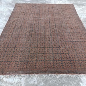 May include: A large, rectangular rug with a repeating floral pattern in dark brown against a lighter brown background. The rug has a fringe along the bottom edge and is placed on a concrete surface.