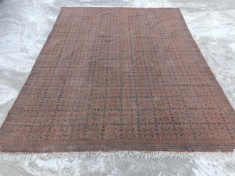 May include: A large, rectangular rug with a repeating floral pattern in dark brown against a lighter brown background. The rug has a fringe along the bottom edge and is placed on a concrete surface.