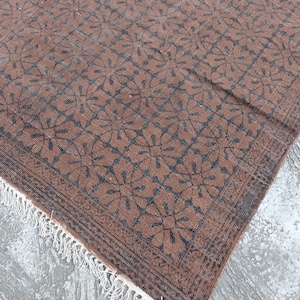 May include: A brown rug with a repeating floral pattern in dark blue. The rug has a decorative border and white fringe along one edge. The rug is made of a woven material and appears to be handmade.
