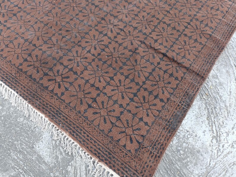 May include: A brown rug with a repeating floral pattern in dark blue. The rug has a decorative border and white fringe along one edge. The rug is made of a woven material and appears to be handmade.