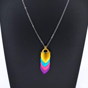 May include: A silver chain necklace with a pendant featuring layered heart-shaped scales in yellow, blue, pink, and purple. The pendant is suspended from a gold-colored bail. The necklace is displayed against a black background.