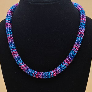 May include: A necklace made of small metal rings in shades of pink, blue, and purple. The necklace is on a black velvet display stand.