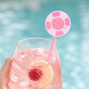 May include: A clear stemless glass filled with a pink beverage, ice cubes, a lemon slice, and a fresh raspberry. A pink straw features a round topper with alternating pink and white segments and the text JetZero Cabana Club in gold lettering.