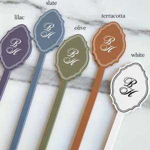 May include: Five long-stemmed markers in lilac, slate, olive, terracotta, and white. Each marker features a scalloped, shield-shaped top with a white or black monogram of the letters BH in a cursive script, surrounded by a thin double-line border.