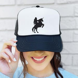 May include: A black and white trucker hat with a silhouette of a person riding a unicorn on the front panel. The hat has a black peak and a black mesh back. A fashionable accessory for everyday wear.