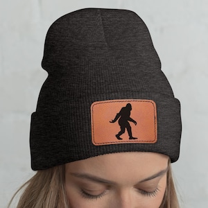 Op de afbeelding: Een donkergrijze beanie met een bruine leren patch met een zwart silhouet van Bigfoot. De beanie is gemaakt van een zacht, gebreid materiaal. De persoon die de hoed draagt, heeft lang blond haar.