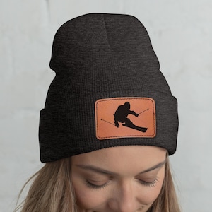 Peut inclure: Bonnet gris foncé avec un patch en cuir marron représentant une silhouette noire d'un skieur. Le bonnet est porté par une personne aux longs cheveux blonds et un haut blanc.