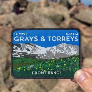 Peut inclure: Un écusson rectangulaire avec une bordure bleue et un motif de paysage de montagne. L'écusson comporte le texte "GRAYS & TORREYS" et "FRONT RANGE", avec des mesures de 14 275 F et 4 351 m. Le motif comprend une chaîne de montagnes, de l'herbe verte et des fleurs jaunes.