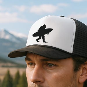 Könnte beinhalten: Schwarz-weiße Trucker-Cap mit einer Silhouette einer Person, die ein Surfbrett trägt. Die Kappe hat einen schwarzen Schirm und eine Netzrückseite mit einer weißen Frontplatte. Die Kappe wird von einer Person in einem blauen Hemd getragen.