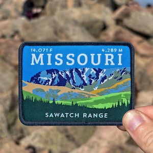 Pode incluir: Um patch retangular com fundo azul exibindo a palavra "MISSOURI" e uma ilustração de uma cordilheira. O patch também inclui o texto "SAWATCH RANGE" e medidas de 14.071 F e 4.289 m. O patch é segurado em frente a um cume rochoso.