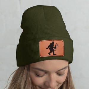 May include: Olive green beanie with a brown leather patch featuring a black silhouette of Bigfoot holding a bow and arrow. The beanie is made of a soft material and has a folded brim.