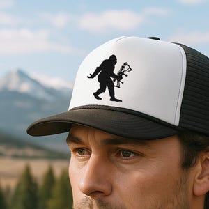 May include: A black and white trucker hat with a Bigfoot silhouette holding a bow and arrow. The hat has a white front panel, a black brim, and a black mesh back. The hat is worn by a person.