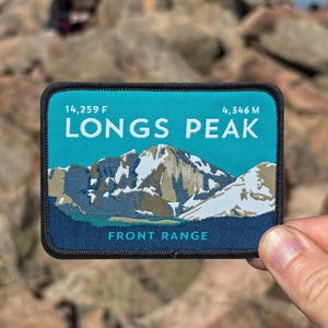 May include: A rectangular patch with a teal background and a mountain illustration. The text "LONGS PEAK" is above the mountain graphic, with "FRONT RANGE" below. The patch also displays the elevation in feet and metres.