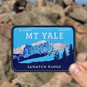 May include: A rectangular patch featuring a mountain landscape with the text "MT YALE" and "SAWATCH RANGE". The patch has a blue background with white and blue mountain imagery and dark blue trees. The patch also includes the text "14,200 F" and "4,328 M".