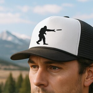 Könnte beinhalten: Schwarz-weiße Trucker-Cap mit einer Silhouette einer Person, die eine Scheibe wirft. Die Kappe hat eine weiße Frontplatte mit der Grafik, einen schwarzen Schirm und eine schwarze Mesh-Rückseite. Die Kappe wird von einer Person getragen.