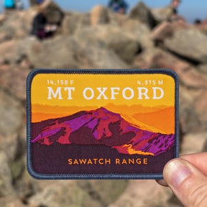 May include: A rectangular patch with a mountain landscape design, displaying "MT OXFORD" and "SAWATCH RANGE". The patch showcases a yellow and orange sky above a purple and red mountain range. It is held in front of a rocky mountain peak.
