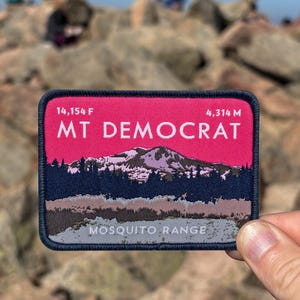 May include: A rectangular patch with a pink background and the text "MT DEMOCRAT" in white. The patch features a mountain range graphic and the text "MOSQUITO RANGE". The patch also includes the elevation in feet and metres.