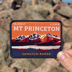 May include: A rectangular patch featuring a mountain range design with the text "MT PRINCETON" and "SAWATCH RANGE". The patch also includes the elevation in feet and meters. The patch is held in front of a rocky mountain landscape.