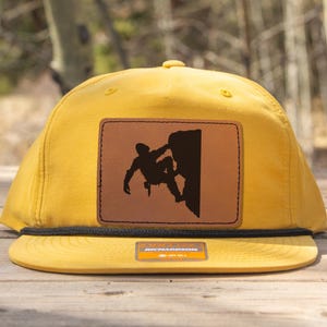 Könnte beinhalten: Senfgelbe Baseballkappe mit flachem Schirm. Ein brauner Lederaufnäher zeigt eine schwarze Silhouette eines Kletterers. Die Kappe hat ein schwarzes Seildetail und ein kleines Etikett auf dem Schirm.