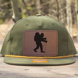 Può includere: Cappellino verde oliva con toppa in pelle marrone raffigurante una silhouette nera di un escursionista con zaino e bastoncino da trekking. Il cappellino ha un dettaglio in corda gialla e una piccola etichetta arancione. Lo sfondo è una vista sfocata di alberi.