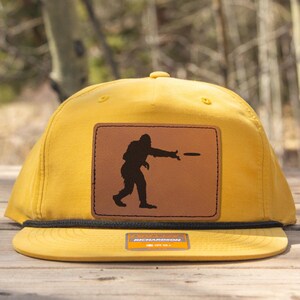 Könnte beinhalten: Senfgelbe Baseballkappe mit schwarzem Schirm und einem braunen Lederaufnäher. Der Aufnäher zeigt eine schwarze Silhouette einer Figur, die eine Scheibe wirft. Ein klassisches, lässiges Accessoire.