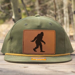 Könnte beinhalten: Olivgrüne Baseballkappe mit einem braunen Lederaufnäher mit einer schwarzen Silhouette von Bigfoot. Die Kappe hat ein gelbes Seildetail und einen flachen Schirm. Der Hintergrund ist eine verschwommene Ansicht von Bäumen.