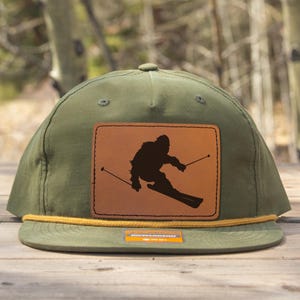 Peut inclure: Casquette vert olive avec un patch en cuir marron représentant une silhouette de skieur noir. La casquette a un détail de corde jaune autour du bord et une petite étiquette. Pour les amateurs de sports d'hiver.