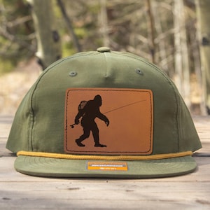 Könnte beinhalten: Olivgrüne Baseballkappe mit einem braunen Lederpatch mit einer schwarzen Silhouette von Bigfoot beim Angeln. Die Kappe hat ein gelbes Seildetail und einen flachen Schirm. Der Hintergrund ist eine verschwommene Ansicht von Bäumen.
