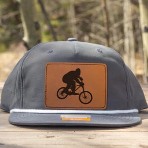 Könnte beinhalten: Graue Baseballkappe mit flachem Schirm und weißem Seildetail. Die Kappe hat einen braunen Lederaufnäher mit einer schwarzen Silhouette eines Mountainbikers auf einem Fahrrad. Der Hintergrund ist eine verschwommene Ansicht von Bäumen.