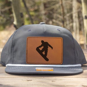 Könnte beinhalten: Graue Flat-Branded-Kappe mit weißem Seildetail und braunem Lederaufnäher. Der Aufnäher zeigt eine schwarze Silhouette eines Snowboarders. Die Kappe liegt auf einer Holzoberfläche.