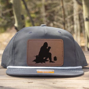 Könnte beinhalten: Graue Baseballkappe mit einem braunen Lederpatch mit einer schwarzen Silhouette einer angelnden Person. Die Kappe hat ein weißes Seildetail und einen flachen Schirm. Der Hintergrund ist eine verschwommene Szene im Freien.