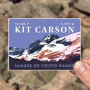 May include: A sticker featuring a stylized illustration of Kit Carson Peak in the Sangre de Cristo Range. The sticker is blue, purple, and white with a snowy mountain peak. The text on the sticker reads "14,165 ft Kit Carson 4,317 m Sangre de Cristo Range".