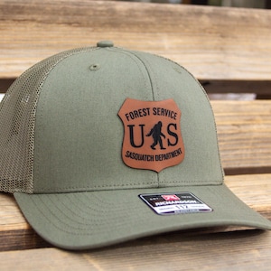 May include: A green and black trucker hat with a brown leather patch that says "Forest Service U.S. Sasquatch Department".